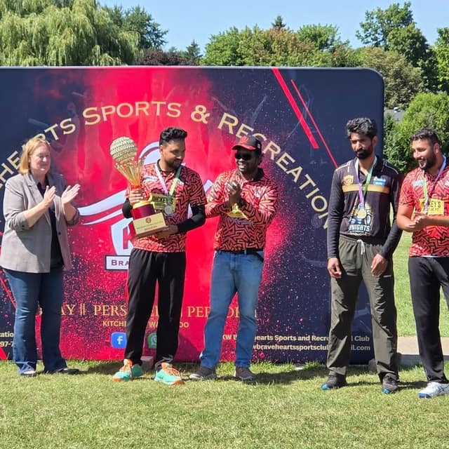 A woman in a gray blazer applauds as a man in a red jersey holds a gold trophy, with two other men beside a Sports & Recreation banner.