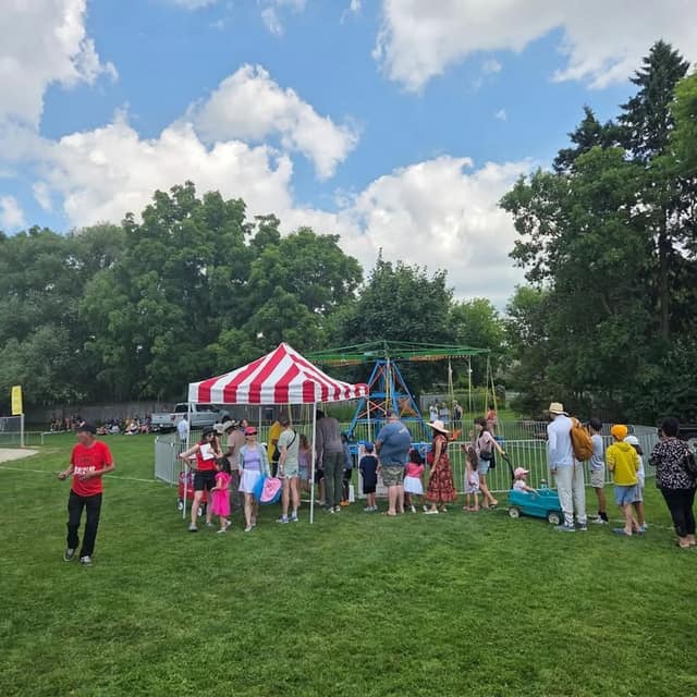 Families and children line up beside a small swing ride and a red and white striped tent on the grass at a summer festival.