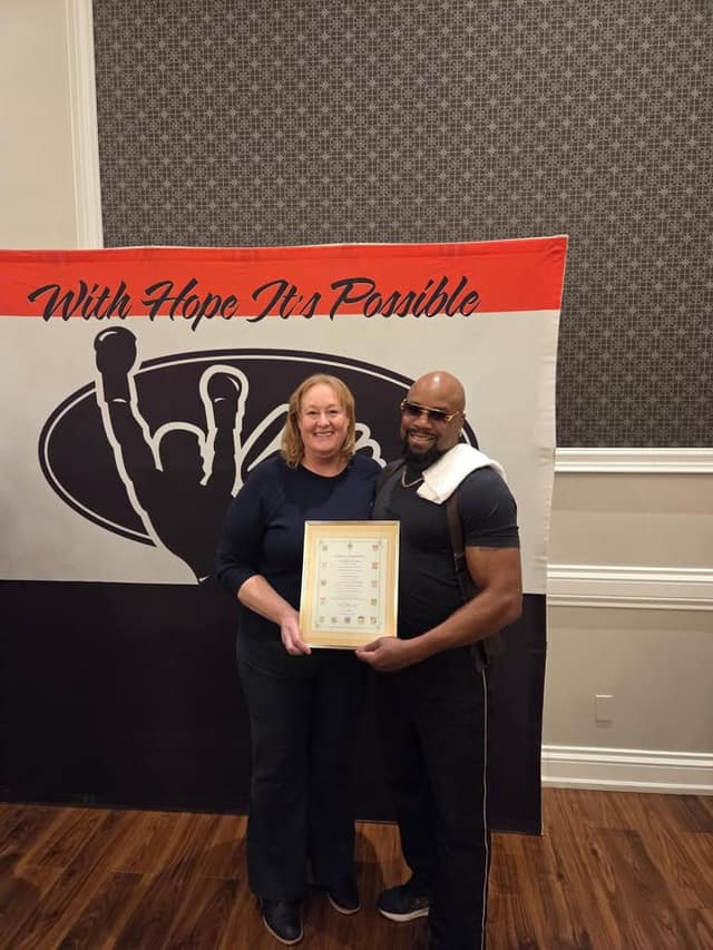 A woman and a man in sunglasses hold a framed certificate in front of a banner reading "With Hope It's Possible".