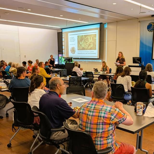 Attendees sit at tables in a meeting room as a woman presents beside a projected slide titled Cultivating Food Sovereignty Together.