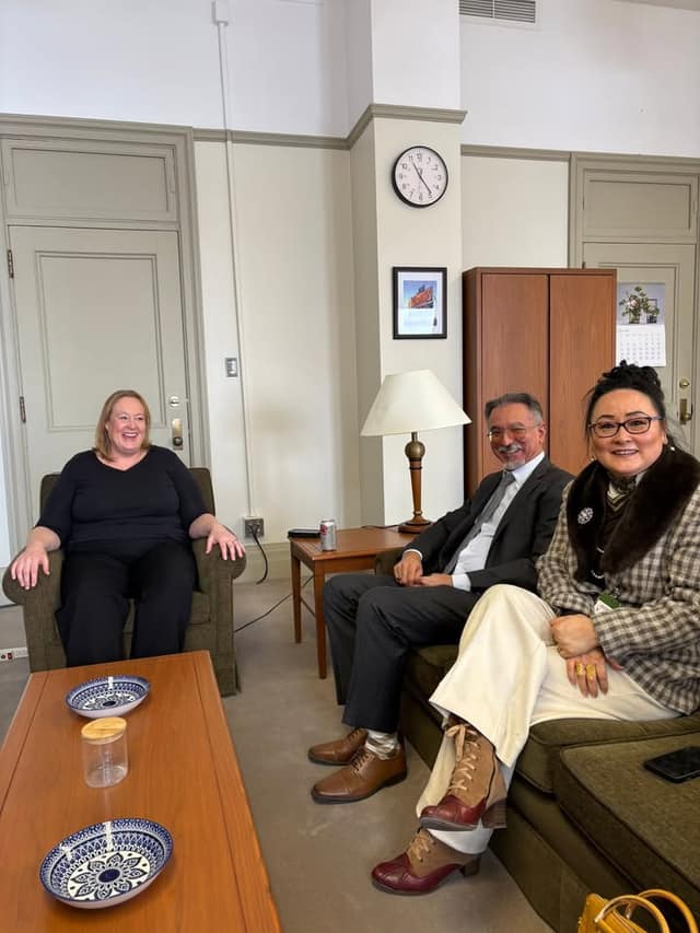 A woman in an armchair laughs while a man in a suit and a woman in a plaid coat sit on a sofa, in an office with a wooden coffee table.
