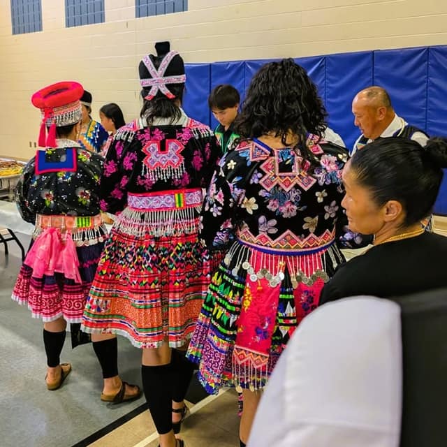 Three women in embroidered black tops and colourful pleated skirts stand in a gym with their backs to the camera.