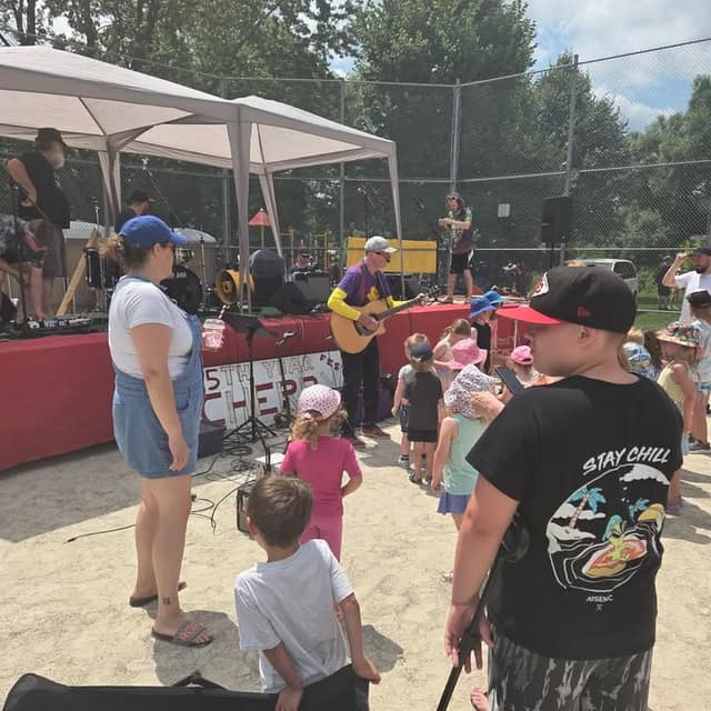 A musician with a guitar performs on a red-skirted stage while toddlers in sun hats and adults watch on a sandy area under a tent.