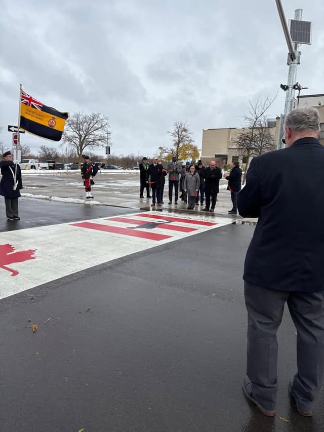 A man in a dark jacket faces a small crowd and a flag bearer beside the painted crosswalk, with a piper standing nearby.
