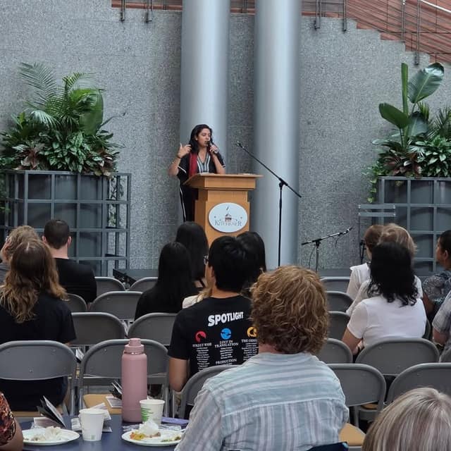 A speaker gestures at a podium with a Kitchener logo while attendees seated in grey chairs listen, one wearing a Spotlight T-shirt.