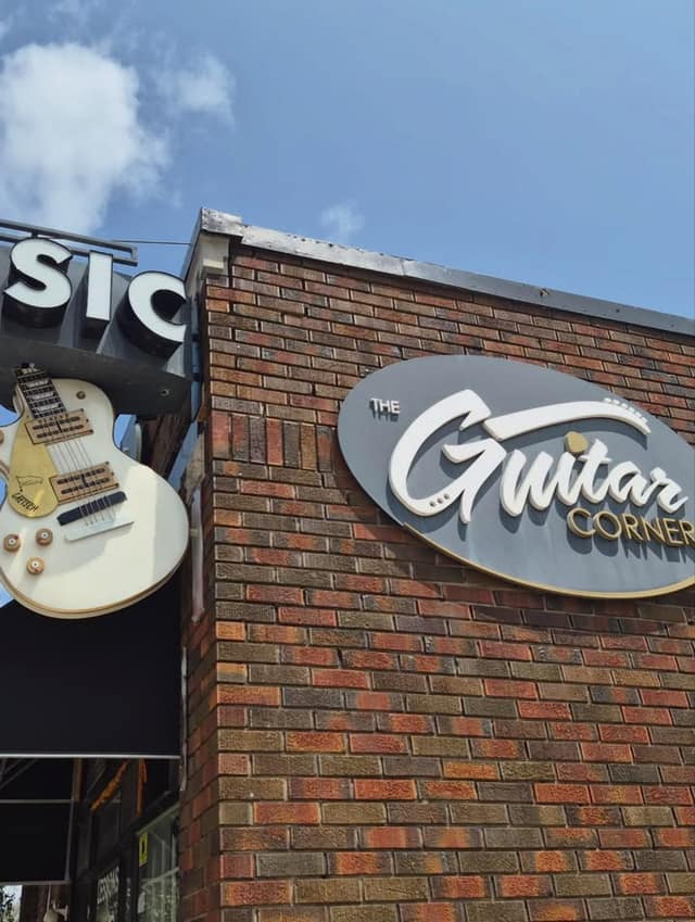 Brick storefront with an oval sign reading The Guitar Corner, and a white electric guitar hanging from a Music sign against a blue sky.
