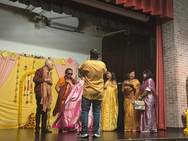 On a stage with yellow and pink drapes and marigold garlands, several women in saris and an older man talk while a person photographs them.