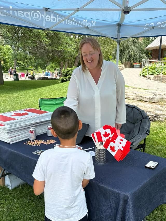 A smiling woman in a white blouse looks at a boy at a table holding Canadian flags, stacked Canada lawn signs and drink cans.