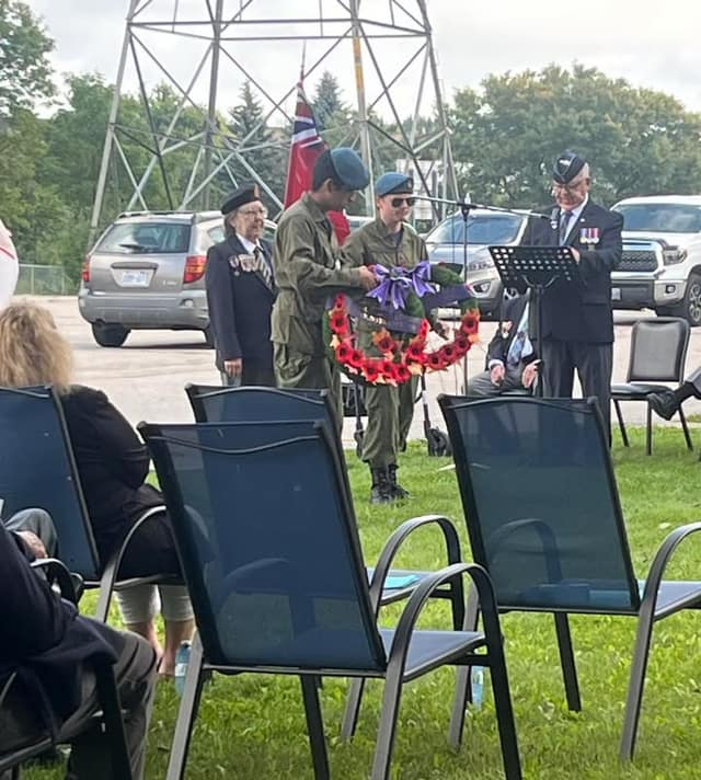 A cadet in green fatigues holds a poppy wreath with purple ribbon as a veteran speaks at a lectern and guests sit nearby.