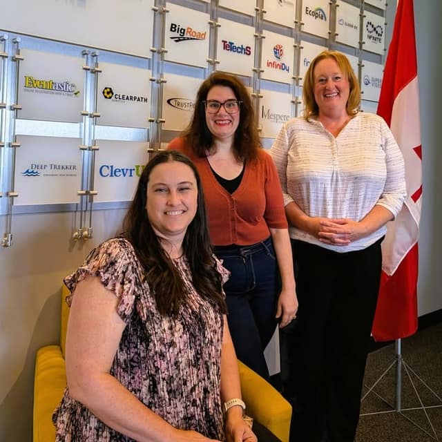 Three smiling women pose in front of a wall of startup company logos beside a Canadian flag; one sits on a yellow chair.