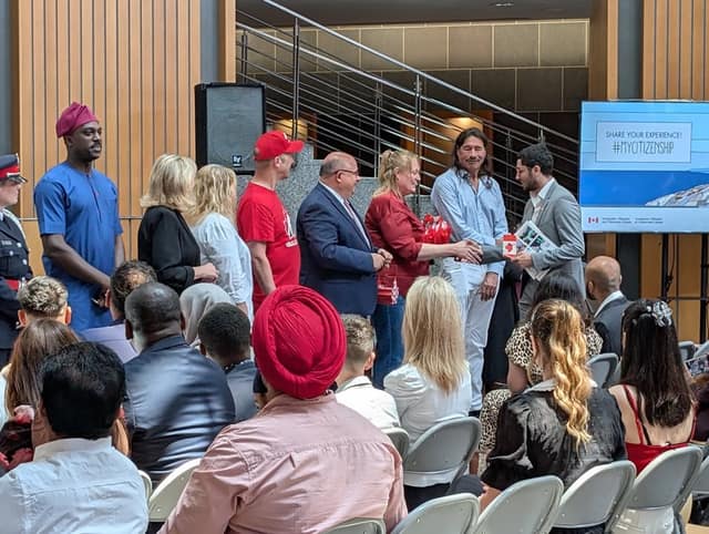 A man in a long-sleeved shirt shakes hands with a woman in red on stage as officials look on, with a #MyCitizenship screen behind them.