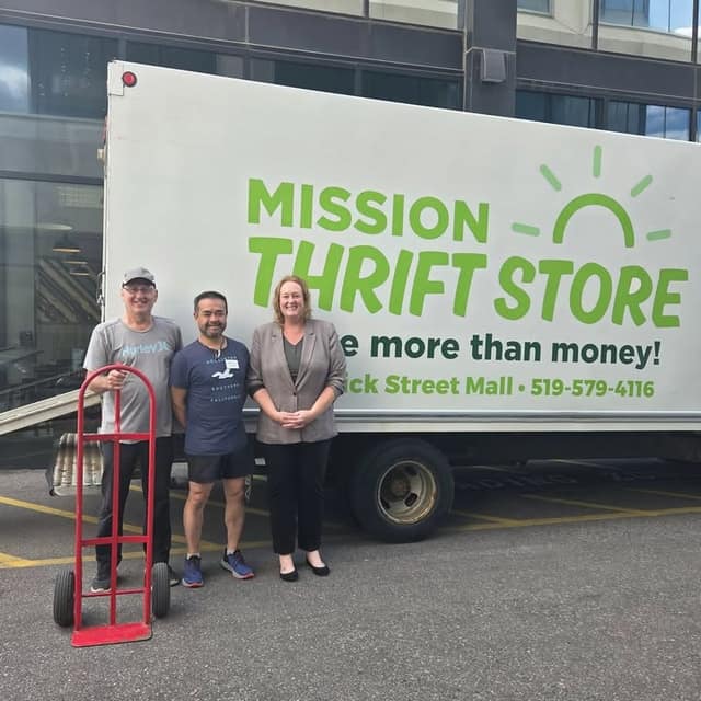 Two men and a woman stand beside a Mission Thrift Store truck in a parking lot, with a red hand truck at left, after a furniture donation.