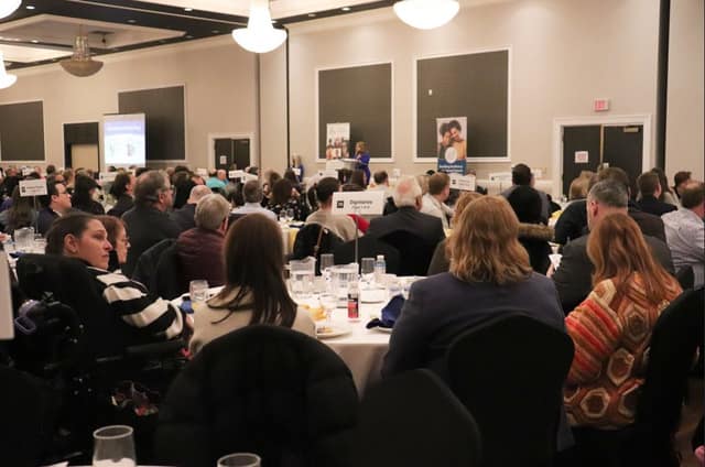 View from behind seated guests at round tables facing a speaker at a podium, with a 'Dignitaries' table sign and a projection screen.