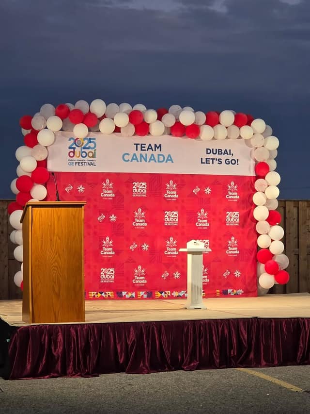 An empty outdoor stage with a wooden podium and balloon arch before a red Team Canada backdrop reading "Dubai, Let's Go!"