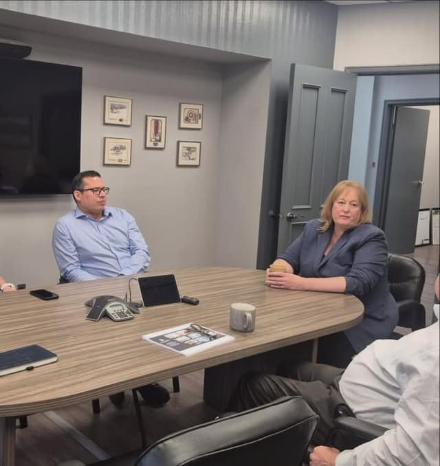 A woman in a blue blazer holding a mug sits at a conference table with a man in glasses and another person; a wall screen is behind them.