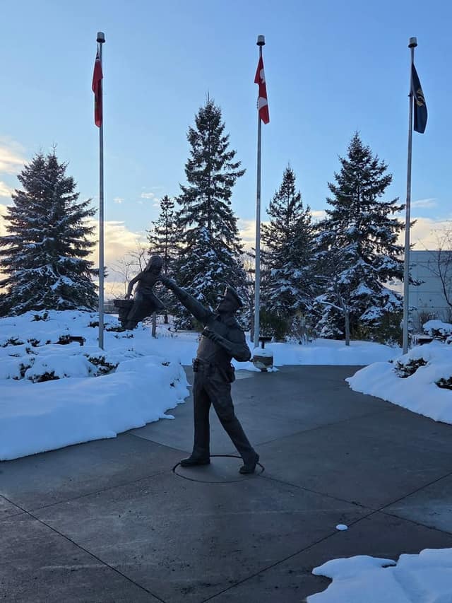 A bronze statue of a police officer swinging a child stands in a snowy plaza, with three flagpoles and snow-covered evergreens behind.