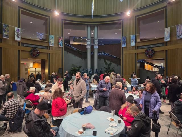 People sit and stand around round tables with cups and donuts in a large indoor hall decorated with wreaths and hanging banners.