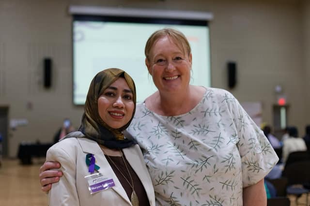 A woman in a hijab and beige blazer with a name tag and ribbon smiles beside a woman in a leaf-patterned blouse in a hall.