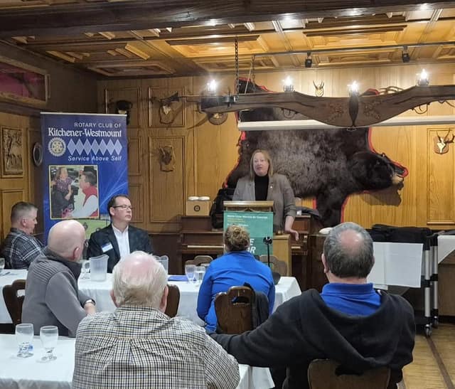 Kelly DeRidder speaks at a podium to seated lunch guests, with a Rotary Club of Kitchener-Westmount banner at left.