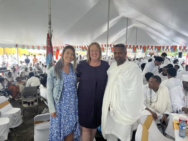 Two women and a man in a white shawl pose together inside the festival tent, with seated guests and Eritrean bunting behind them.