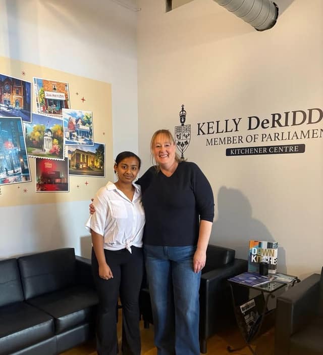 Setna and a woman stand arm in arm in a lobby with black sofas, a postcard mural, and a wall sign reading Kelly DeRidder, Member of Parliament.