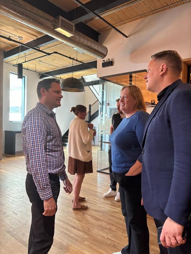 A man in a checked shirt talks with a woman in a blue top and a man in a navy blazer in a bright loft office with wood floors.