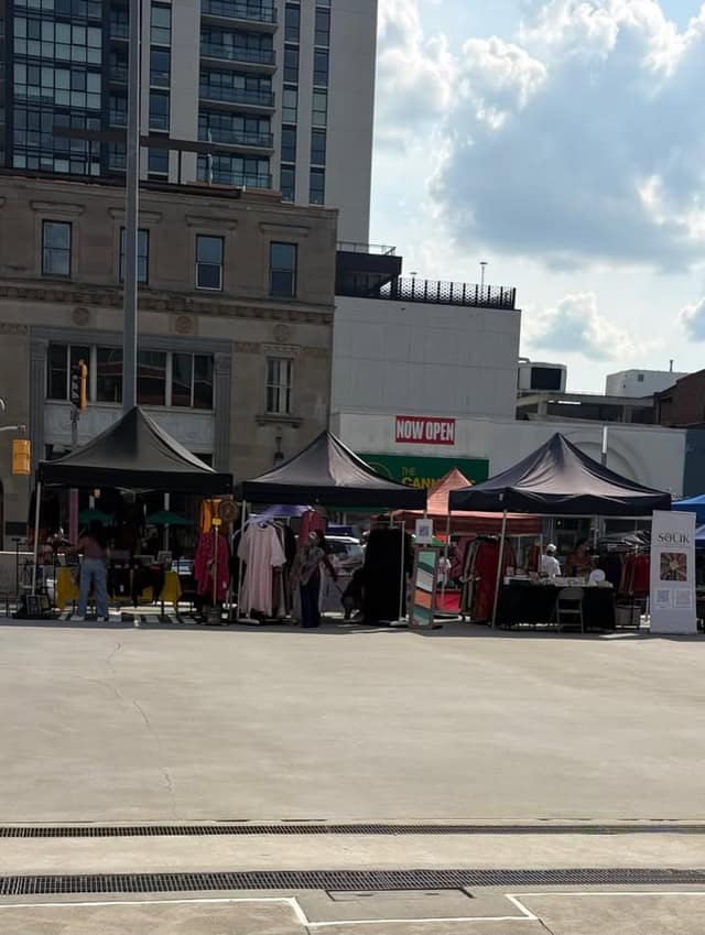 Black vendor tents with racks of clothing line a paved square, with a Souk banner and a Now Open sign on a building behind.
