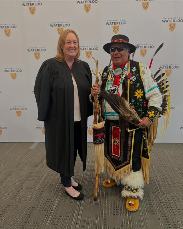 A woman in a black gown stands beside a man in Indigenous regalia with a feathered fan and staff, before a Waterloo backdrop
