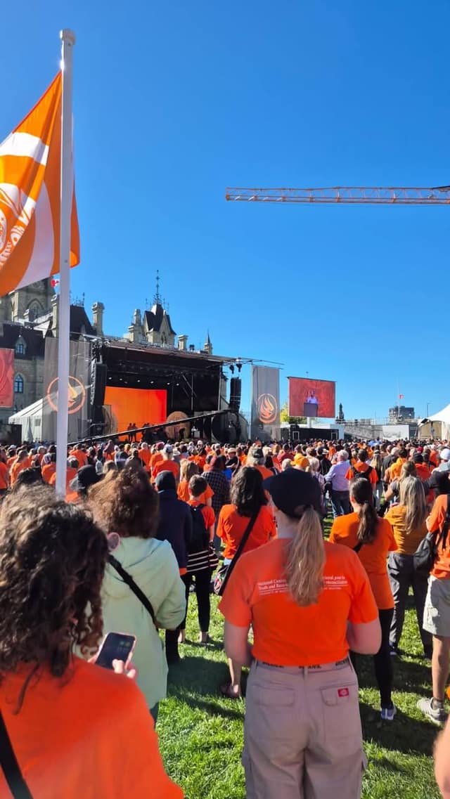 Crowd members in orange shirts stand on the lawn facing a stage and screen, with an orange flag at left and a crane overhead.