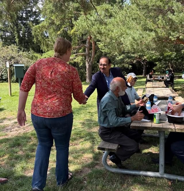 A woman in a red patterned top shakes hands with a smiling man in a navy blazer beside seated people eating at a park picnic table.