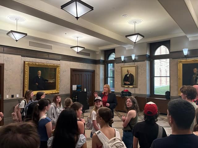 A woman in a dark blazer and red top talks with a group of students in a hall with framed portraits and tall windows.