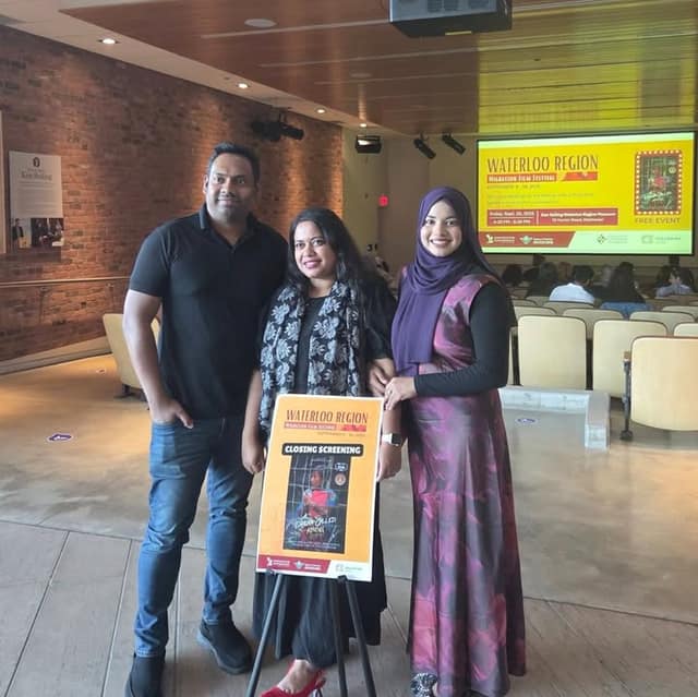 Two women and a man stand beside a Closing Screening easel poster in a hall, with a Waterloo Region Migration Film Festival slide and seated audience behind.
