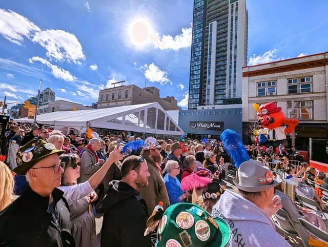 A large crowd in festive hats watches a stage on a sunny downtown street, with a tall building and a red mascot figure behind them.