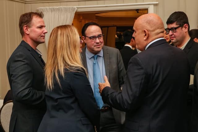 Four men and a woman in business attire stand in a close conversation at an indoor event, with other guests behind them.