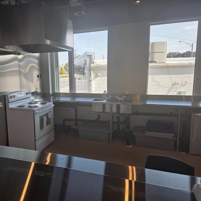 A commercial kitchen with a white electric stove, stainless steel counters and sinks, and large windows facing the street.