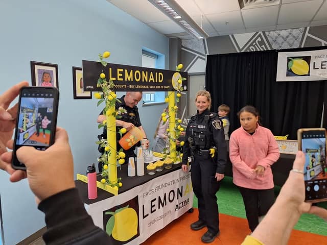 A police officer pours lemonade at a LemonAID stand as another officer and a girl in pink pose while people photograph them.