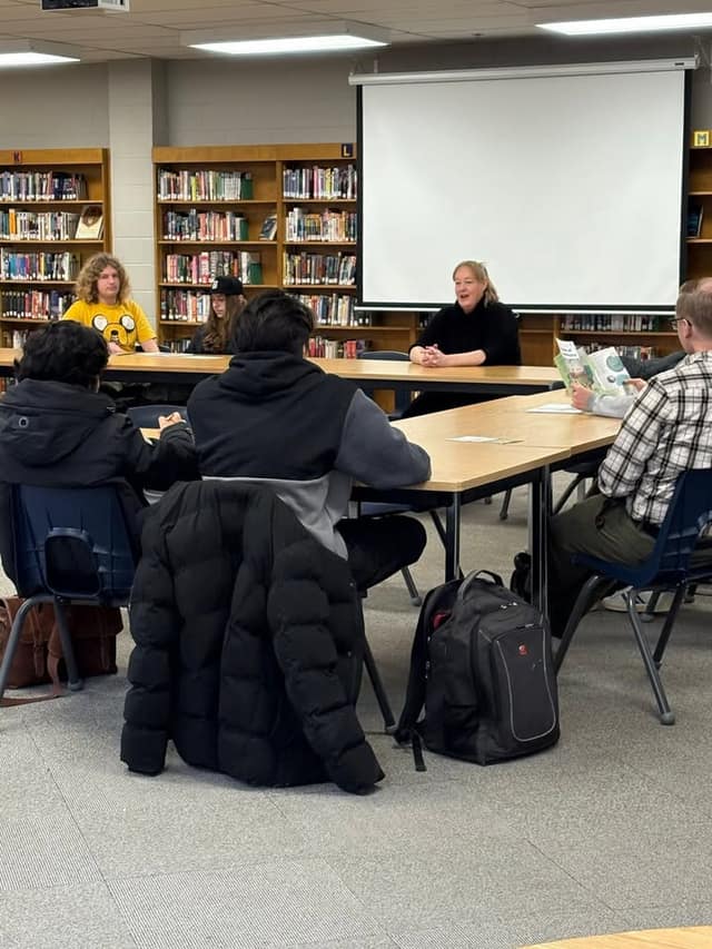 A woman in black speaks to seated students in a school library, with a projection screen behind her and a student holding a brochure.