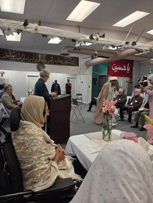 A speaker stands at a wooden podium in a decorated hall as seated guests, including a woman in a wheelchair, and a photographer look on.