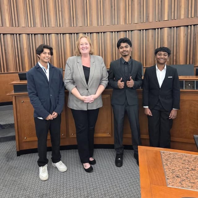 A woman in a gray blazer stands with three smiling young men in suits in a wood-paneled council chamber; one gives thumbs up.