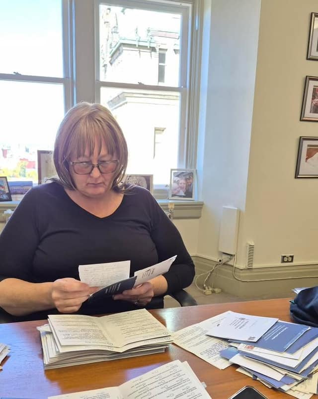 A woman with glasses and shoulder-length hair, in a black top, sits at a wooden desk reading a mailer, with stacks of mail and framed photos around her.