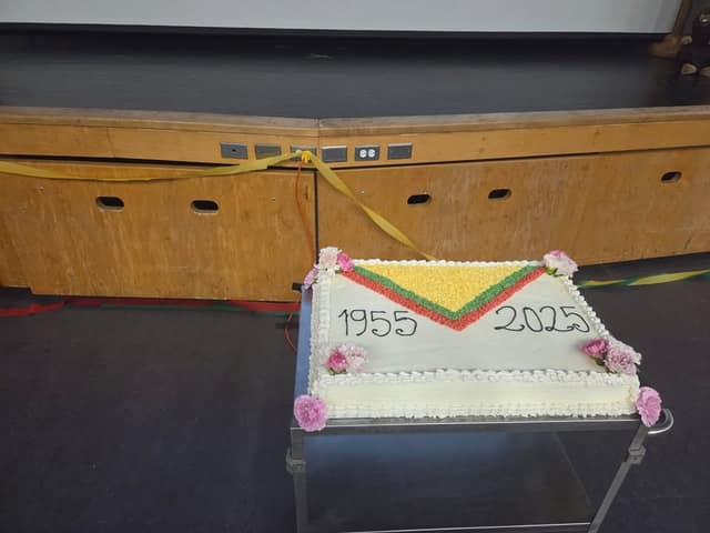 A rectangular white cake with flowers at the corners, a green, yellow and red neckerchief design, and the years 1955 and 2025 sits on a cart.