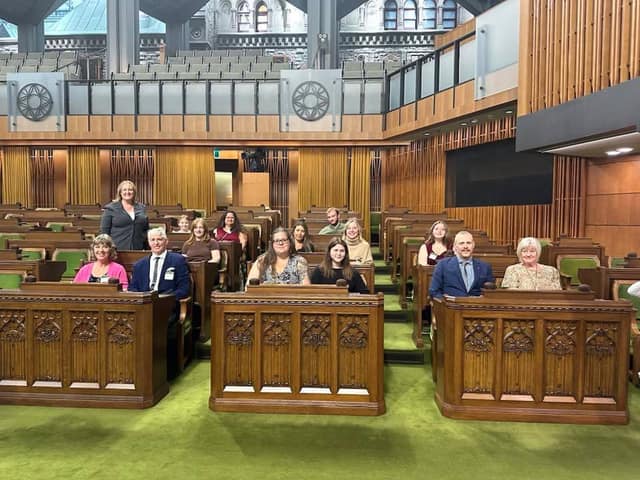 A group of adults sits at wooden desks in the House of Commons chamber with green carpet, while the woman in a gray blazer stands behind them.