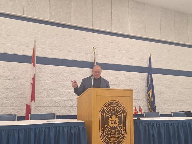 A man in a gray jacket and glasses points while speaking at the Local 527 podium, with Canadian and union flags behind him.