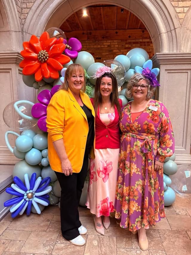 Three smiling women pose together in front of a pastel balloon arch with large flower balloons; two wear floral fascinators.
