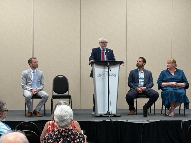 A man in a navy suit and red tie speaks at a podium reading "Bingemans" as three panelists sit nearby and audience members watch.