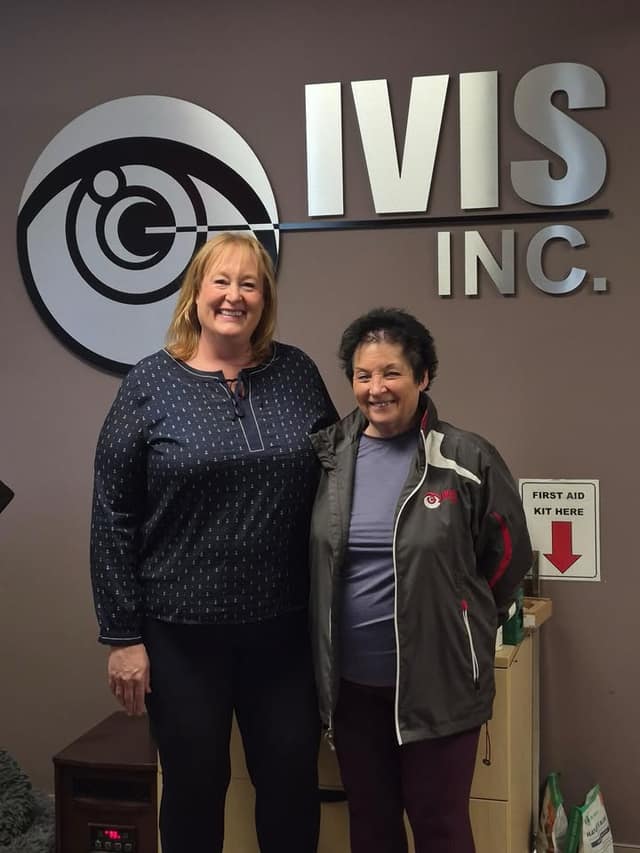 Two smiling women stand side by side in front of a wall sign reading "IVIS INC." with an eye logo; a first aid kit sign is at right.