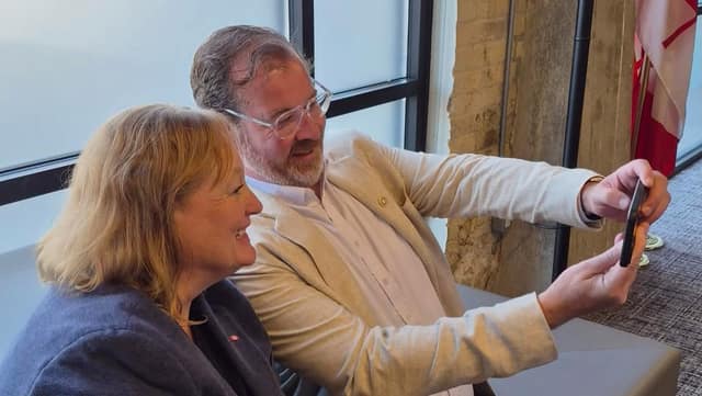 A woman with blonde hair smiles as a bearded man in clear glasses and a beige blazer holds up a phone to take a selfie with her.