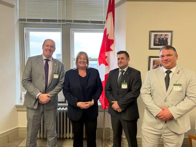 Three men in suits and a woman in a dark blazer smile in an office beside a Canadian flag, with visitor badges on their jackets.
