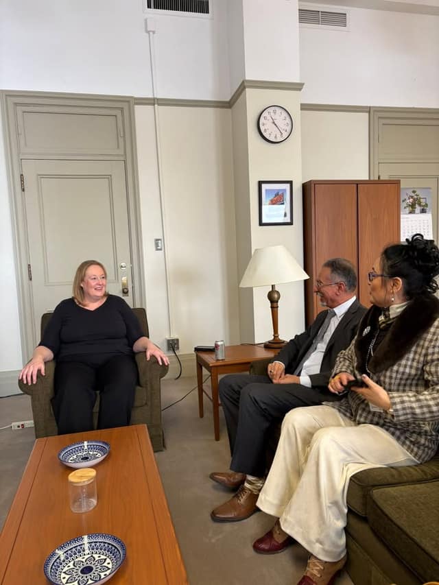 A woman in an armchair talks with a man in a suit and a woman in a plaid coat seated on a sofa, in an office with a clock on the wall.