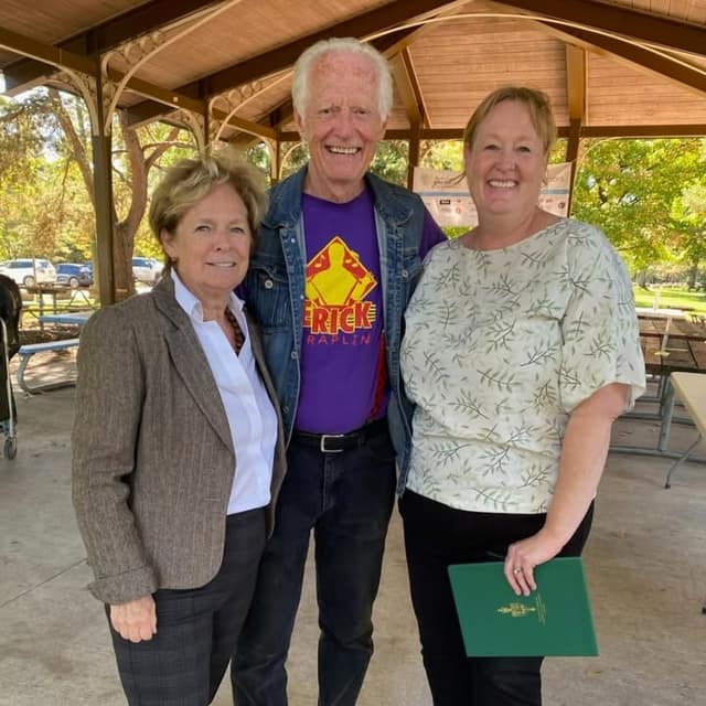 Three smiling people pose together under a park pavilion; one wears a purple T-shirt and denim jacket, another holds a green folder.